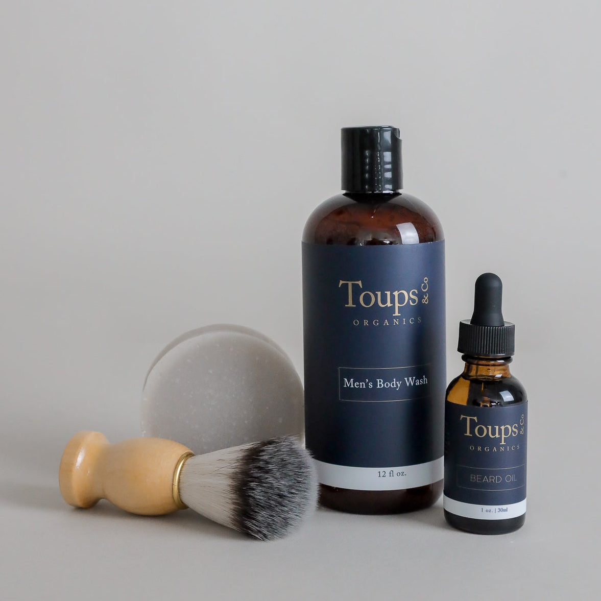 The Men’s Grooming Set by Toups and Co Organics, featuring a brush, round soap bar, a brown bottle of body wash, and a dropper bottle of beard oil—all branded with Toups and Co Organics—is arranged on a neutral background, ideal for natural men’s grooming and skincare routines.