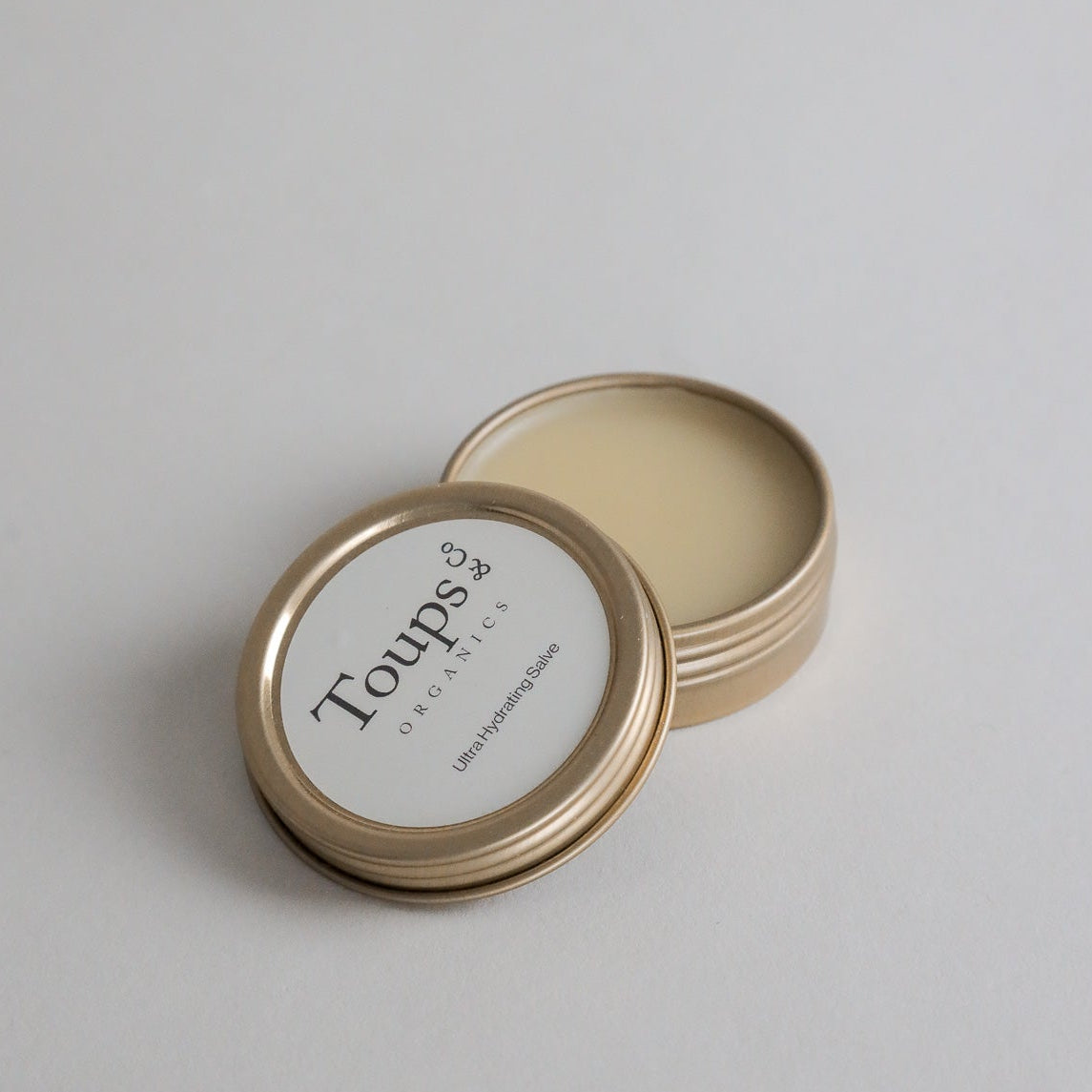 A small open tin of Toups and Co Organics Ultra Hydrating Salve rests on a plain white surface, revealing the pale, deeply moisturizing salve inside—an ideal choice for any all-natural skincare routine.