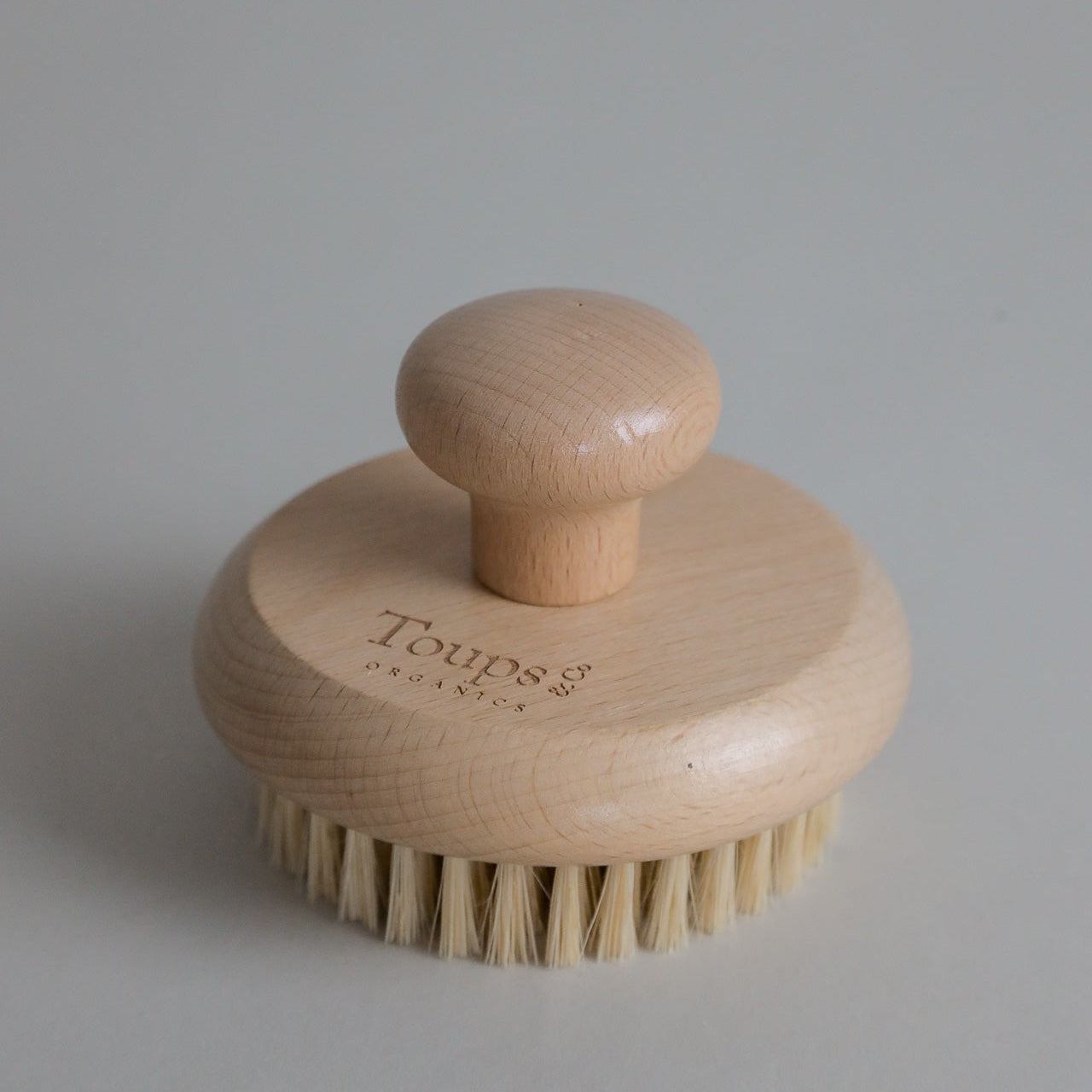 A Dry Brush by Toups and Co Organics, featuring a round wooden body with a short handle and natural sisal bristles, engraved with “Toups” on the top—perfect for dry brushing and supporting lymphatic drainage, shown on a plain light background.
