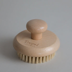 A Dry Brush by Toups and Co Organics, featuring a round wooden body with a short handle and natural sisal bristles, engraved with “Toups” on the top—perfect for dry brushing and supporting lymphatic drainage, shown on a plain light background.