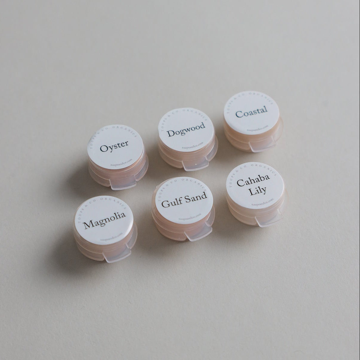 Six small round containers from the Toups and Co Organics Sample Set - Liquid Foundation, labeled Dogwood, Magnolia, Coastal, Cahaba Lily, Oyster, and Gulf Sand, are arranged on a grey surface—ideal for sampling foundation shades.