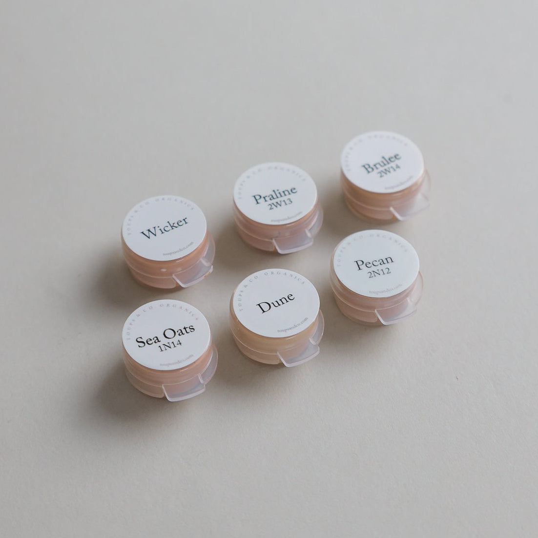 Six small round containers from the Toups and Co Organics Sample Set - Liquid Foundation, labeled Wicker, Praline, Brulee, Sea Oats, Dune, and Pecan, are arranged on a beige surface—ideal for sampling foundation shades.