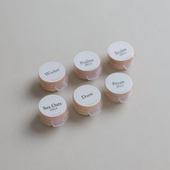 Six small round containers from the Toups and Co Organics Sample Set - Liquid Foundation, labeled Wicker, Praline, Brulee, Sea Oats, Dune, and Pecan, are arranged on a beige surface—ideal for sampling foundation shades.
