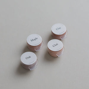Four small round containers from the Toups and Co Organics Sample Set - Liquid Foundation, labeled Maple, Clay, Oak, and Chai are arranged on a beige surface—ideal for sampling foundation shades. 