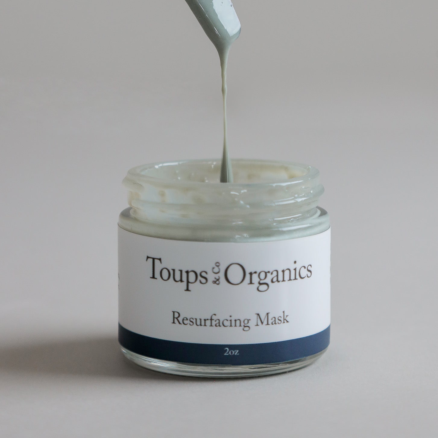 A jar labeled Toups and Co Organics Resurfacing Mask, with a spatula lifting light green cream from the open container, enriched with blue algae extract for powerful anti-aging benefits.