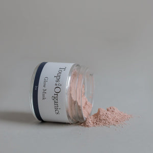 A glass jar labeled Toups and Co Organics Glow Face Mask lies on its side, spilling a pile of Australian pink clay powder onto a light gray surface.
