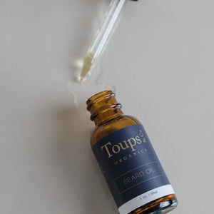 A brown glass bottle of Toups and Co Organics Beard Oil sits open on a surface, its dropper resting beside it and a small pool of oil spilled nearby, showcasing this nourishing beard oil—an essential moisturizer crafted to support healthy beard growth.