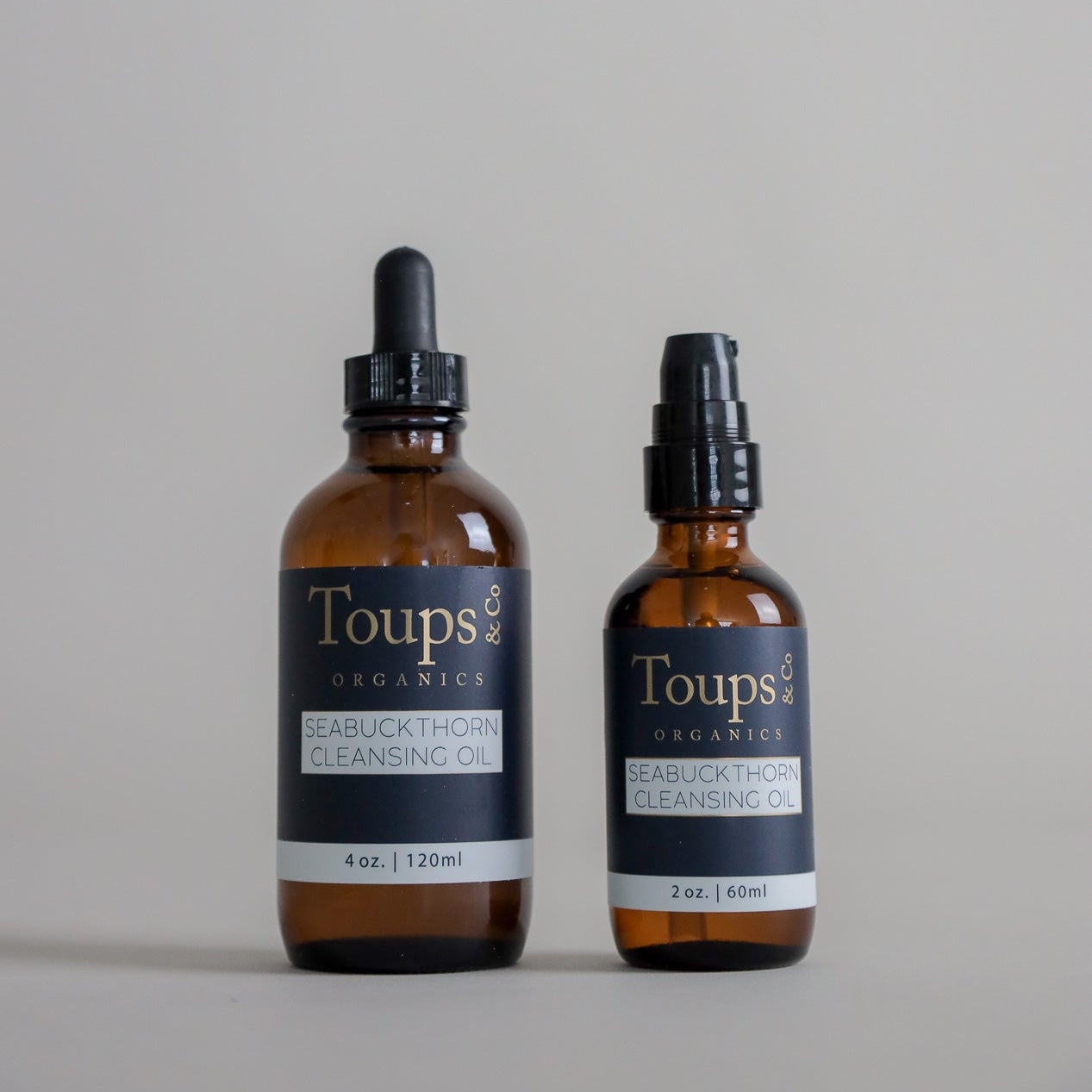 Two amber bottles of Toups and Co Organics Seabuckthorn Cleansing Oil—a nourishing cleanser—are displayed on a plain background. The 4 oz (120 ml) bottle features a dropper, while the 2 oz (60 ml) bottle comes with a pump, providing versatile options for those seeking an effective oil cleanser and gentle makeup remover.