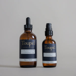 Two amber bottles of Toups and Co Organics Seabuckthorn Cleansing Oil—a nourishing cleanser—are displayed on a plain background. The 4 oz (120 ml) bottle features a dropper, while the 2 oz (60 ml) bottle comes with a pump, providing versatile options for those seeking an effective oil cleanser and gentle makeup remover.