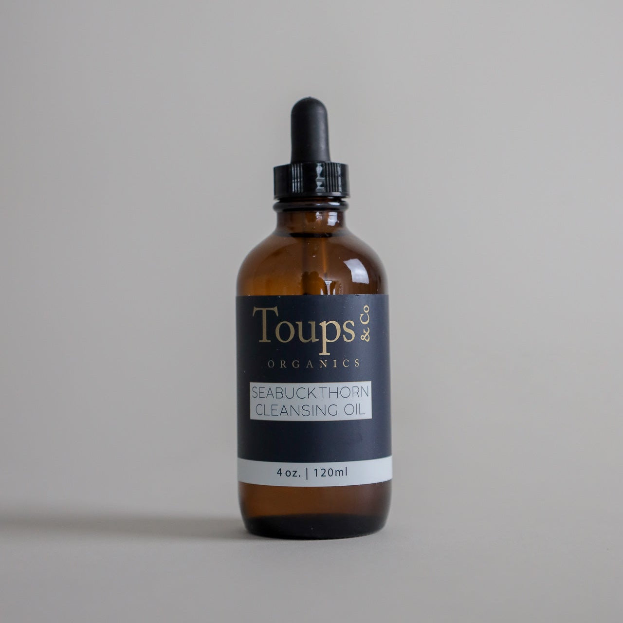 A 4 oz (120 ml) brown glass bottle of Toups and Co Organics Seabuckthorn Cleansing Oil, featuring a black dropper cap, is displayed against a plain gray background. This nourishing cleanser gently purifies the skin and removes makeup, helping maintain your skin’s natural balance.