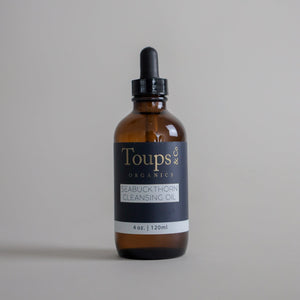 A 4 oz (120 ml) brown glass bottle of Toups and Co Organics Seabuckthorn Cleansing Oil, featuring a black dropper cap, is displayed against a plain gray background. This nourishing cleanser gently purifies the skin and removes makeup, helping maintain your skin’s natural balance.