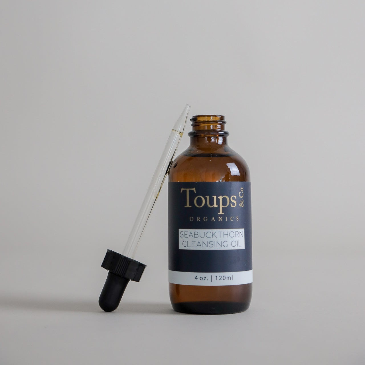 A brown glass bottle labeled “Seabuckthorn Cleansing Oil” by Toups and Co Organics, a nourishing cleanser and gentle makeup remover, with a dropper resting against it on a plain light background.