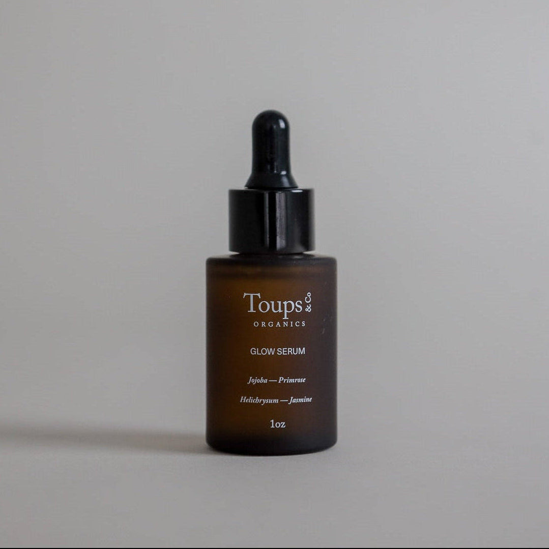 A brown glass dropper bottle labeled “Glow Serum” from Toups and Co Organics, made with vitamin-rich oils for radiant skin, is set against a plain gray background. The label clearly displays the list of ingredients.