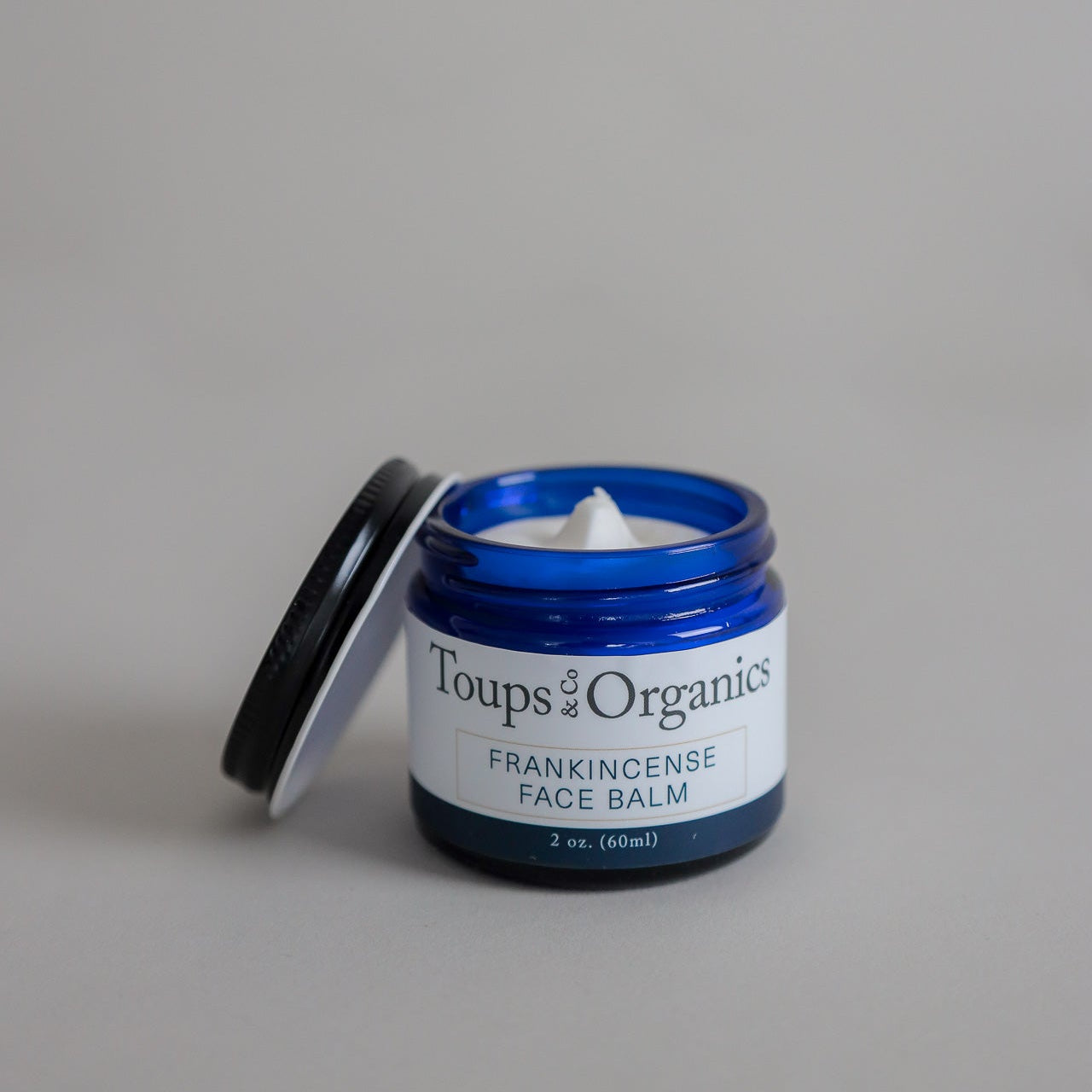 A blue jar labeled Toups and Co Organics Frankincense Face Balm, with its lid removed to display the white cream inside, rests on a simple light background. This nourishing tallow moisturizer is formulated to soothe delicate skin and minimize redness.