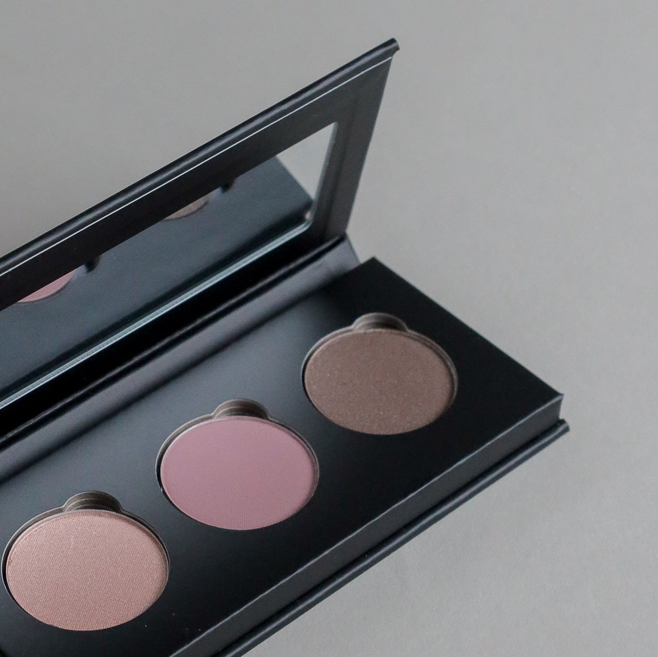 Close-up product photo of a three-shade eyeshadow palette displayed on a clean grey seamless background, showcasing versatile eyeshadow colors and a modern beauty aesthetic.
