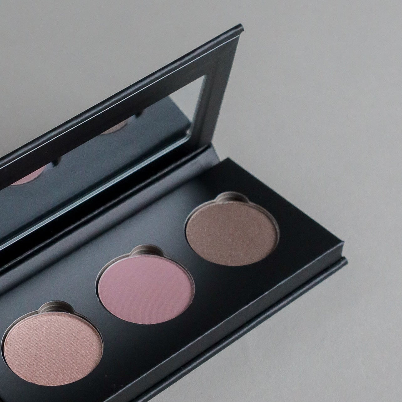Close-up product photo of a three-shade eyeshadow palette displayed on a clean grey seamless background, showcasing versatile eyeshadow colors and a modern beauty aesthetic.