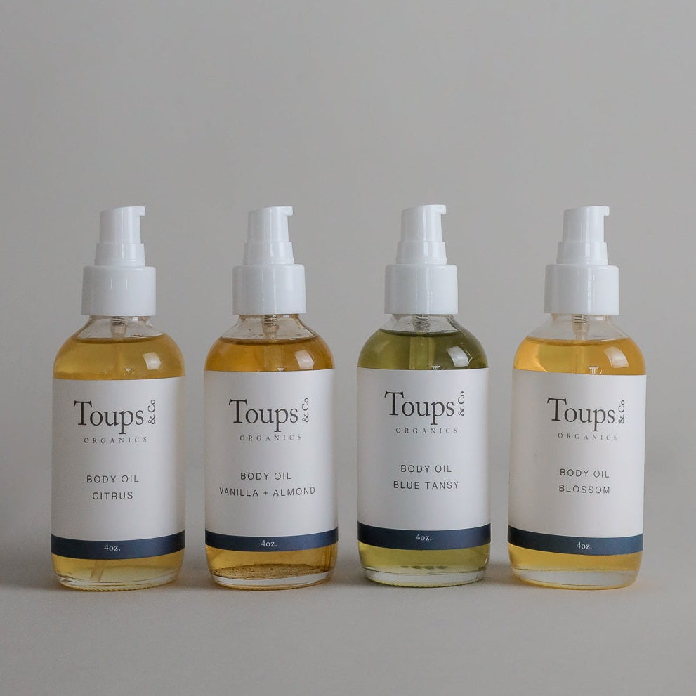 Four 4oz bottles of Toups and Co Organics Organic Body Oil with pump tops in citrus, vanilla almond, blue tansy, and blossom scents, arranged in a row against a neutral background. This lightweight Organic Body Oil nourishes your skin while supporting its natural balance.