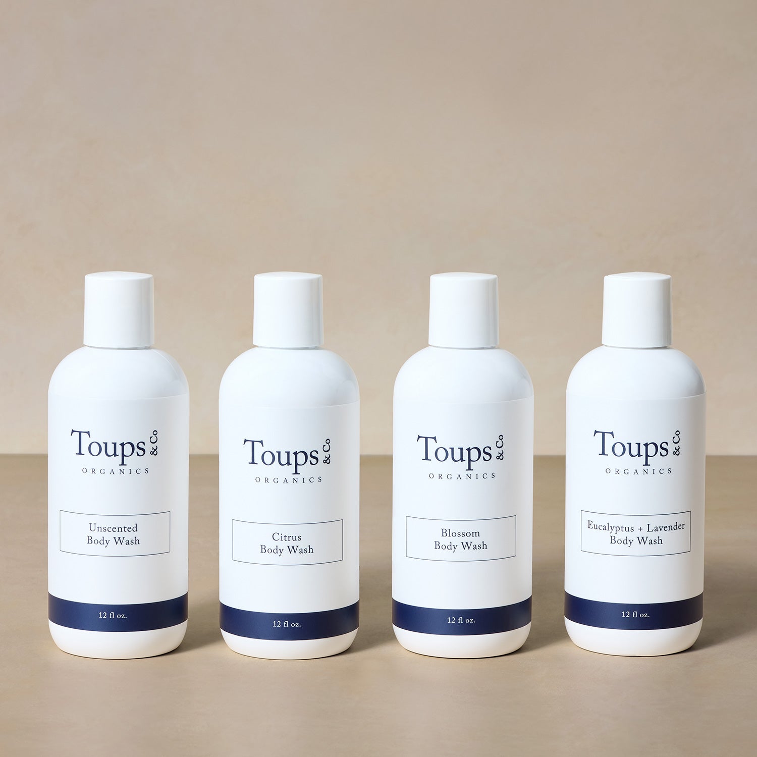Four bottles of Toups and Co Organics Body Wash—Unscented, Citrus, Blossom, and Eucalyptus + Lavender—are lined up side by side against a neutral background.