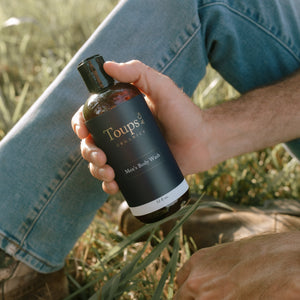 A person sitting on grass holds a bottle labeled Toups and Co Organics Men’s Non Toxic Essentials in their hand, with blue jeans and brown shoes visible.