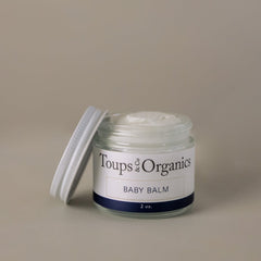 A small glass jar labeled Toups and Co Organics Baby Balm, celebrated for its gentle diaper rash and eczema relief, sits on a plain beige background with the lid resting at an angle to show the white balm inside.
