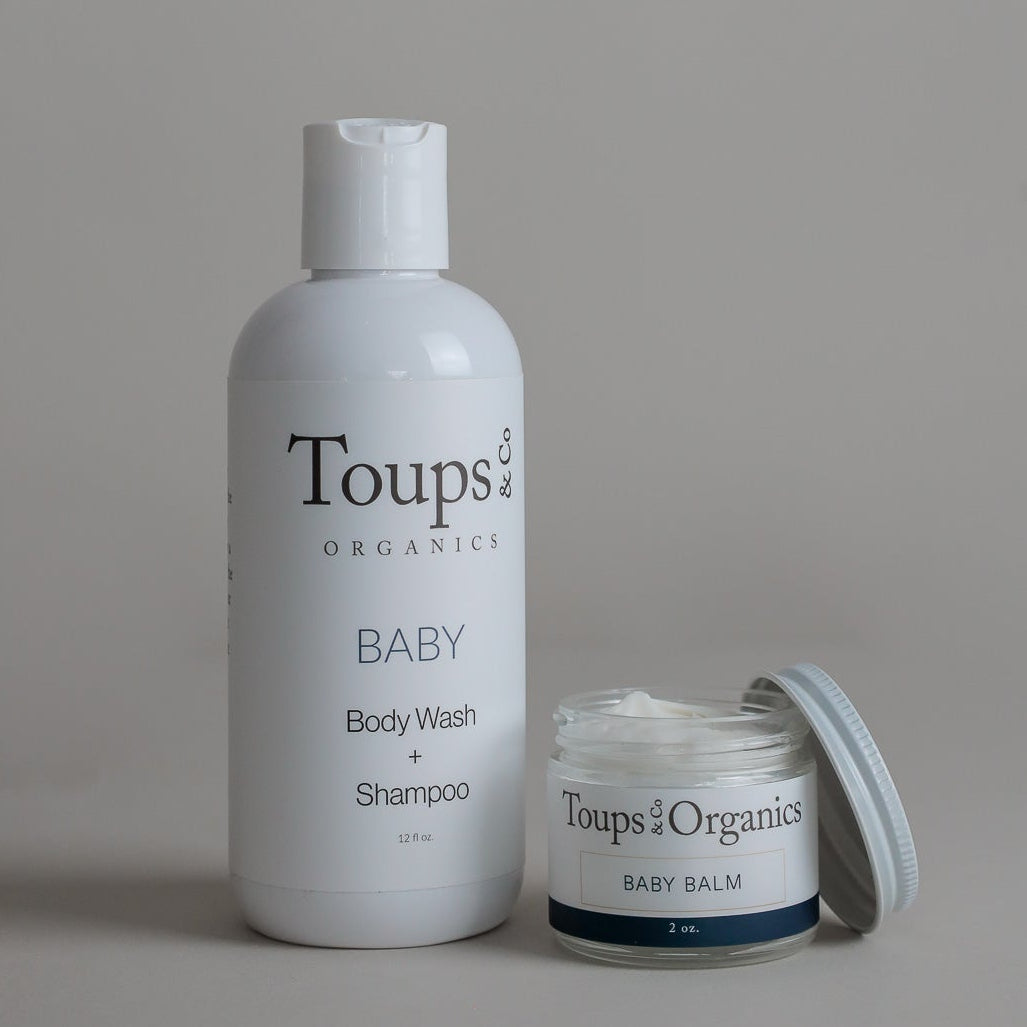 A white bottle from the Toups and Co Organics Baby Essentials Set, ideal for sensitive skin, stands beside an open container from the same set against a plain gray background.