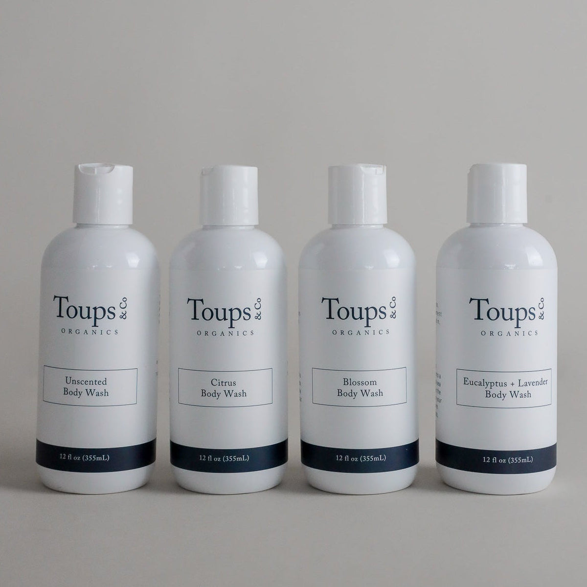 Four white bottles of Toups and Co Organics Body Wash are neatly arranged in a row, each featuring a unique scent: Unscented, Citrus, Blossom, and Eucalyptus + Lavender. Specially formulated for sensitive skin, this hormone-safe and restorative body wash offers a gentle yet luxurious cleansing experience.