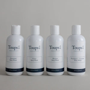 Four white bottles of Toups and Co Organics Body Wash are neatly arranged in a row, each featuring a unique scent: Unscented, Citrus, Blossom, and Eucalyptus + Lavender. Specially formulated for sensitive skin, this hormone-safe and restorative body wash offers a gentle yet luxurious cleansing experience.