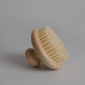 The Toups and Co Organics Dry Brush, featuring a round wooden body, light sisal bristles, and an easy-grip knob handle for effective dry brushing and lymphatic drainage, is displayed upright against a neutral background.