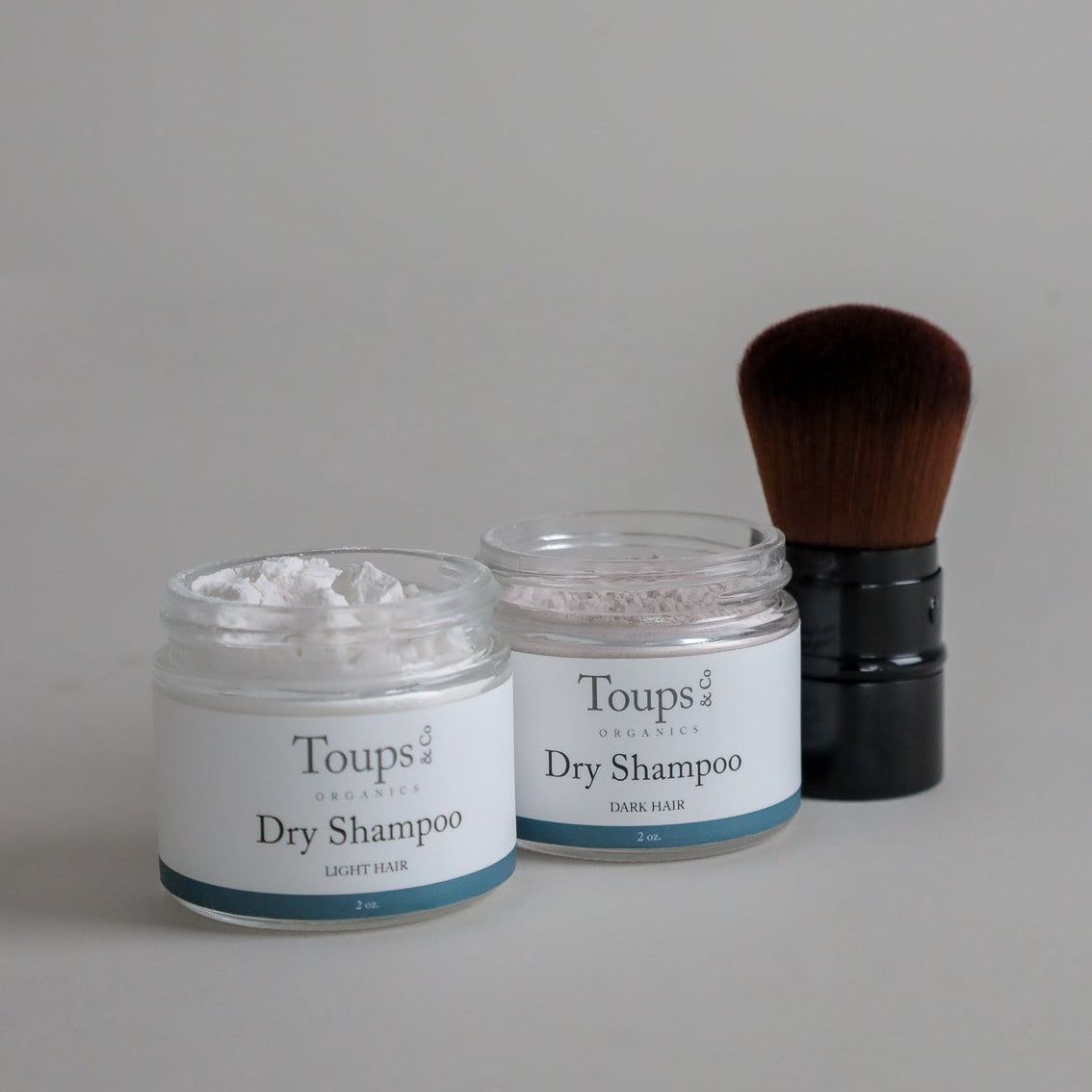 The Toups and Co Organics Clean Hair Set, featuring dry shampoo for both light and dark hair, is displayed beside a large makeup brush on a neutral background—ideal for oil-absorbing freshness and a gentle scalp detox.