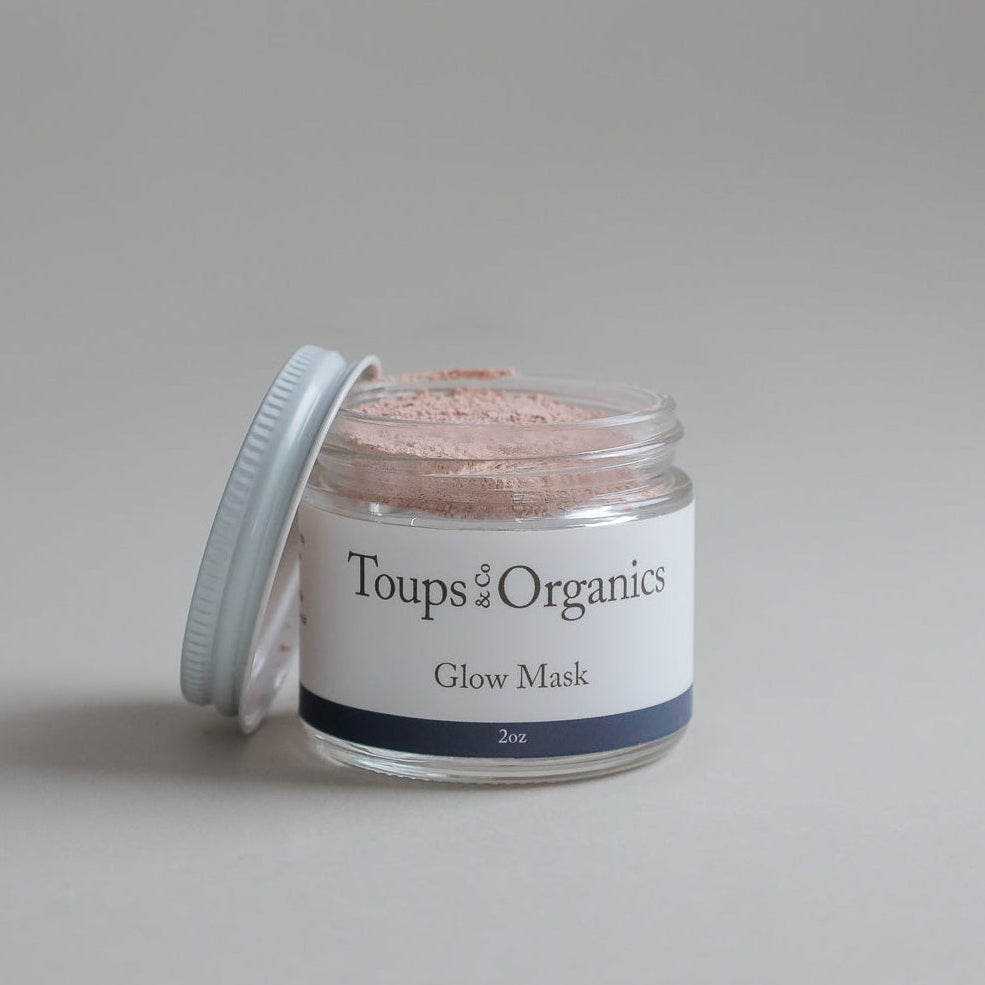 A clear jar labeled Toups and Co Organics Glow Face Mask (2oz) with a white lid, partially open to reveal pink powder inside, is set against a plain light background—ideal for anyone looking for a detoxifying clay mask designed for acne-prone skin.