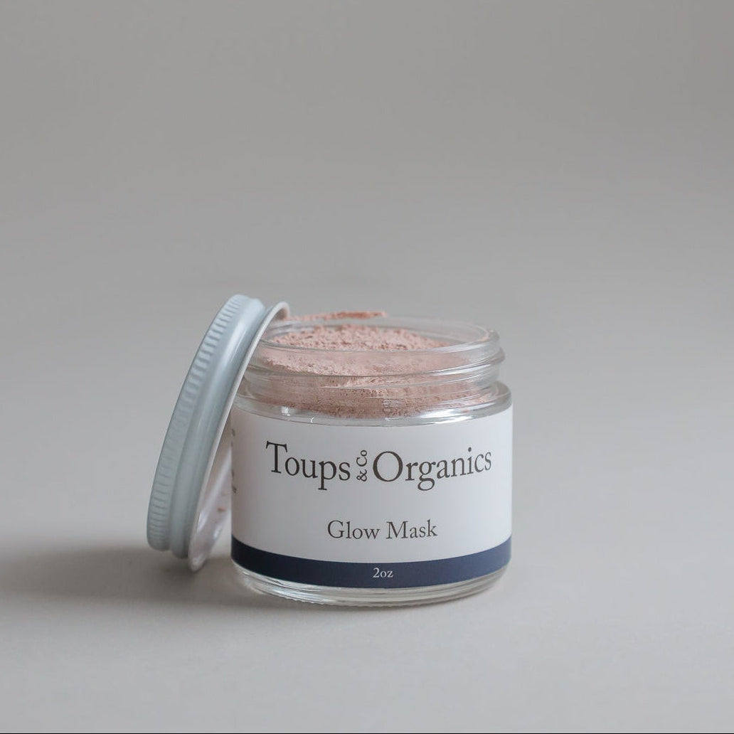 An open glass jar labeled Toups and Co Organics Glow Face Mask sits with its white lid resting nearby, displaying a pink powder made with Australian pink clay inside.