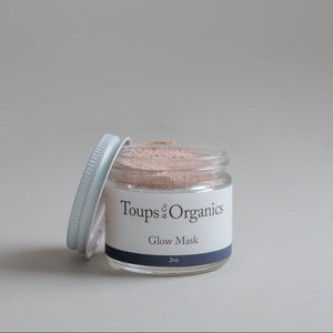An open glass jar labeled Toups and Co Organics Glow Face Mask sits with its white lid resting nearby, displaying a pink powder made with Australian pink clay inside.