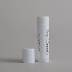 A white tube labeled Hydrating Tallow Stick from Toups and Co Organics, made with nourishing grass-fed tallow, stands upright with its cap placed nearby on a plain light gray background.