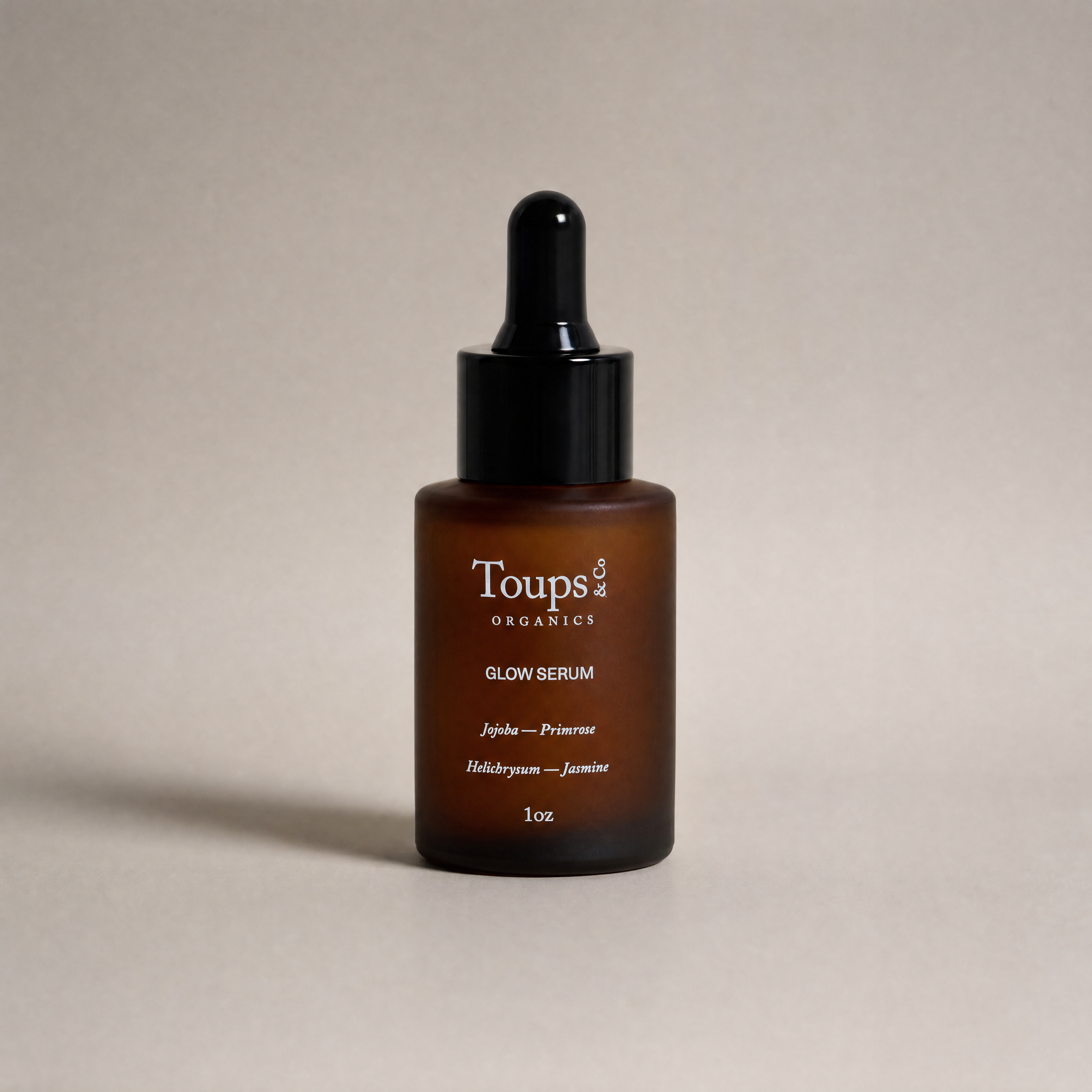 A 1oz brown glass dropper bottle labeled “Glow Serum” by Toups and Co Organics, highlighting vitamin-rich oils and ingredients, displayed against a plain beige background.