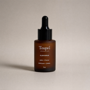 A 1oz brown glass dropper bottle labeled “Glow Serum” by Toups and Co Organics, highlighting vitamin-rich oils and ingredients, displayed against a plain beige background.