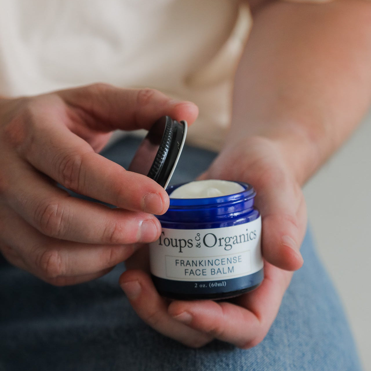 A person opens a blue jar of Toups and Co Organics Men’s Non Toxic Essentials, holding the lid in one hand and the jar in the other.