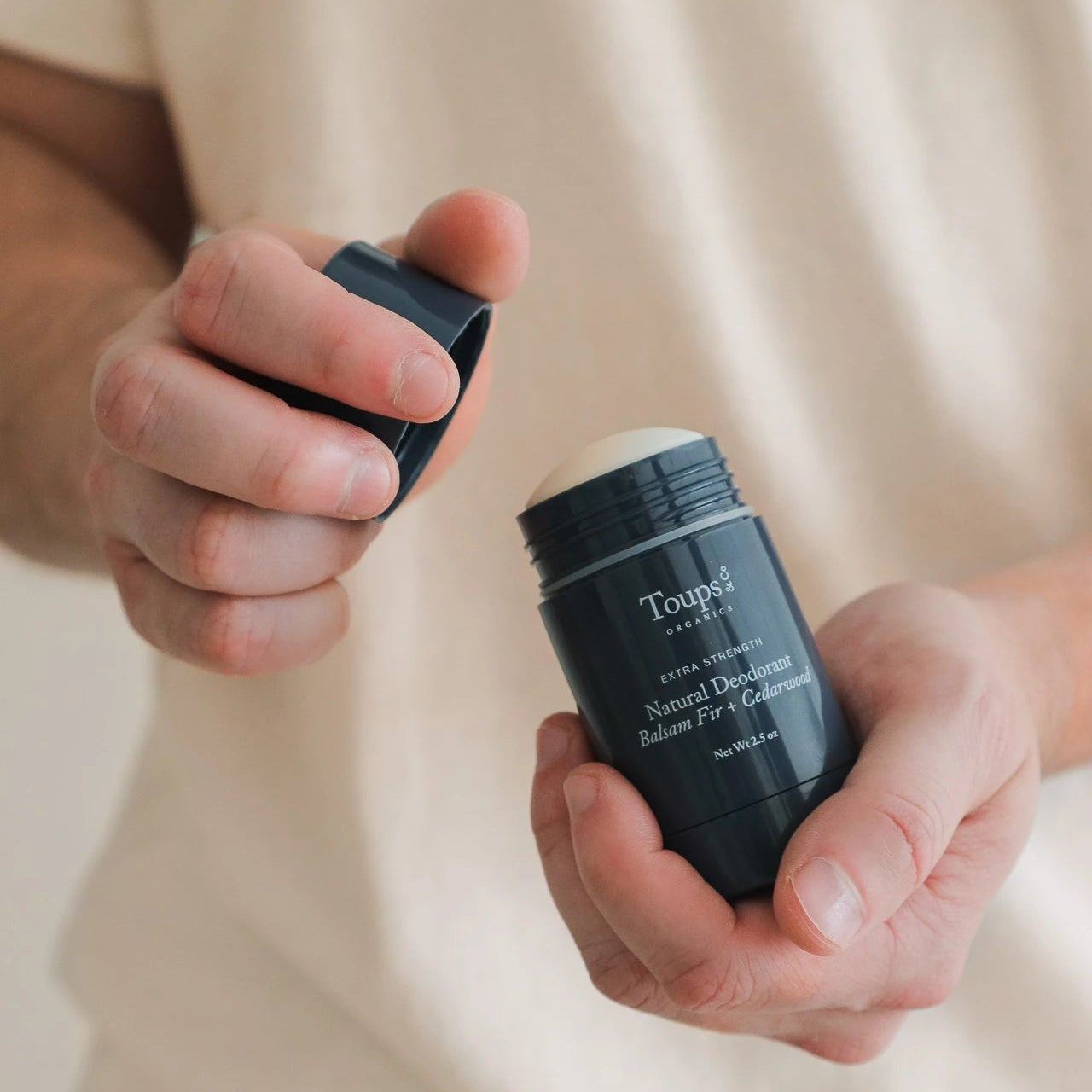 A person holds an open stick of Toups and Co Organics Men's Basics Bundle natural deodorant balm in one hand and the cap in the other.