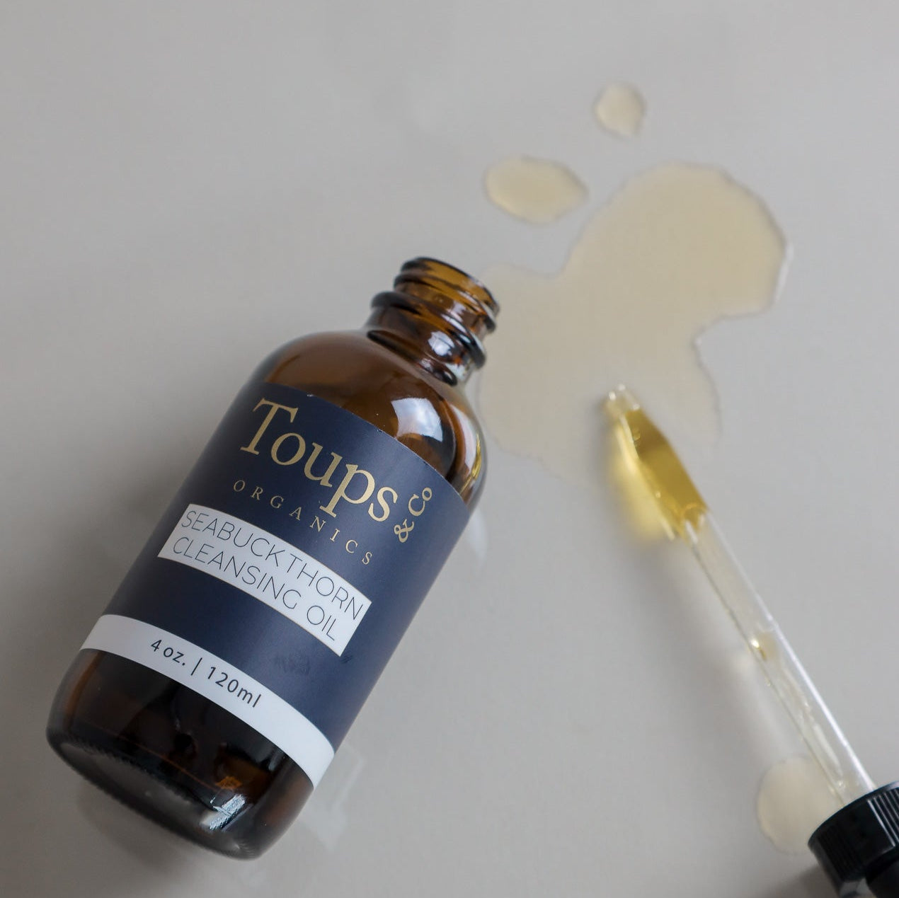 A brown glass bottle labeled Toups and Co Organics Seabuckthorn Cleansing Oil rests on its side with oil spilled beside a dropper on a light surface, emphasizing its role as an effective nourishing skincare oil cleanser and gentle makeup remover.