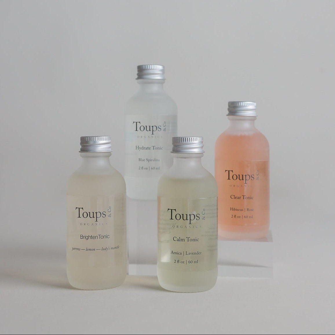 Four labeled glass bottles of Toups and Co Organics Facial Tonic in assorted pastel hues are arranged on a white surface against a minimal background, highlighting their infusion of organic herbs for a revitalizing herbal mist experience.