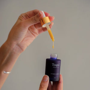 A person holds a dropper filled with yellow serum above a small dark bottle labeled Toups and Co Organics Vitamin C Serum.