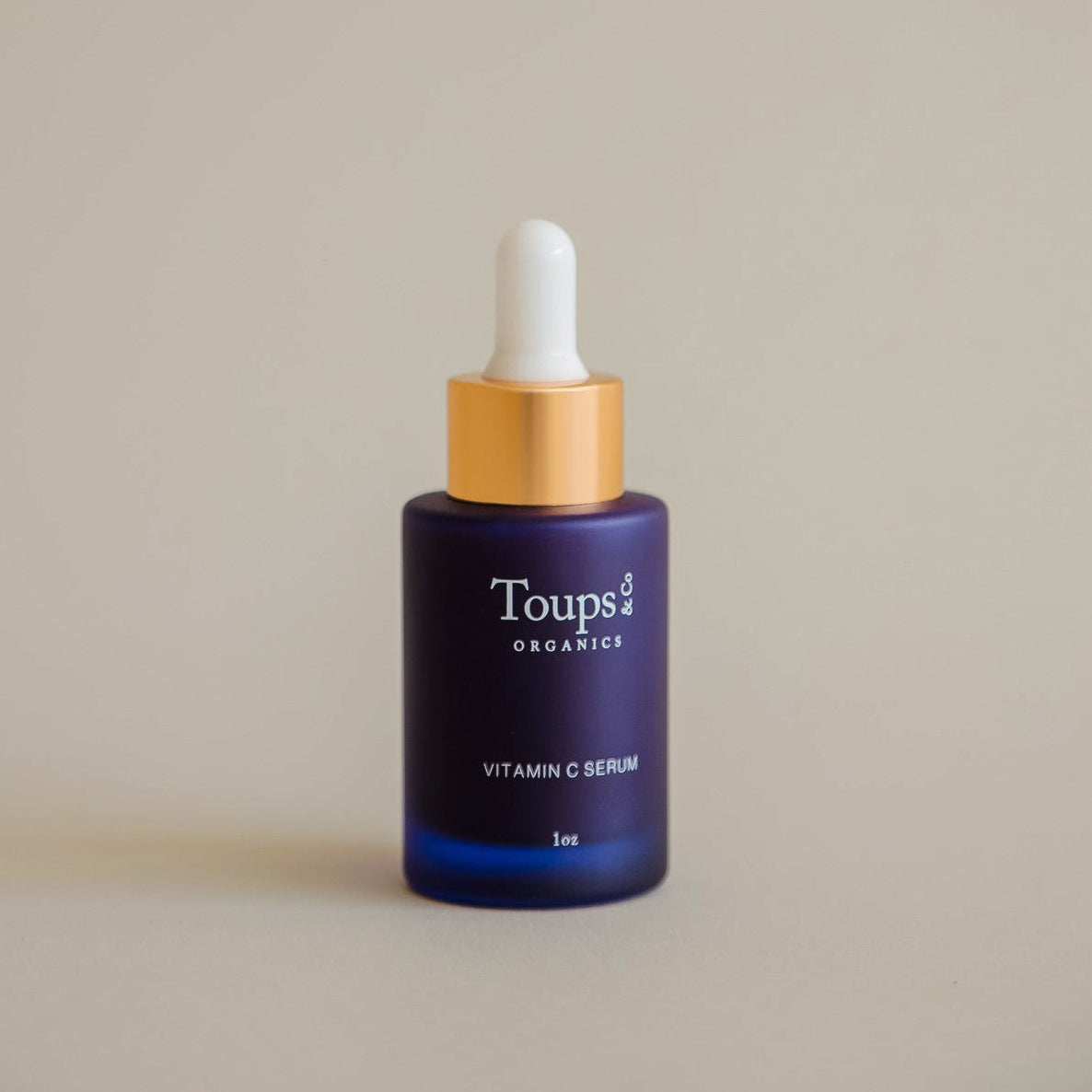 A dark blue dropper bottle labeled Toups and Co Organics Vitamin C Serum, 1oz stands upright against a plain beige background, highlighting its antioxidant formula designed to brighten dull skin.