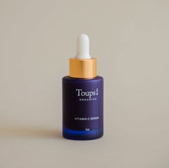 A dark blue dropper bottle labeled Toups and Co Organics Vitamin C Serum, 1oz stands upright against a plain beige background, highlighting its antioxidant formula designed to brighten dull skin.