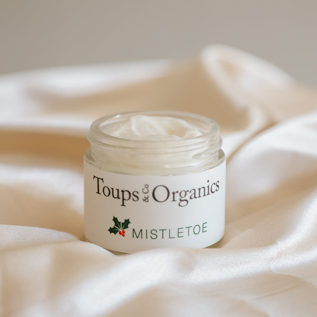 A glass jar of Toups and Co Organics Mistletoe Balm, filled with grass-fed tallow cream, rests on a soft beige fabric—an ideal Christmas gift.