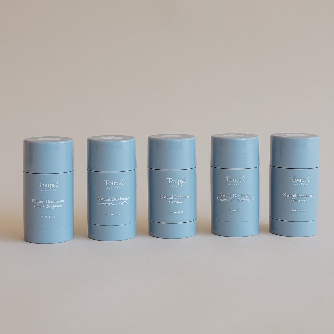 Five light blue cylindrical Toups and Co Organics Natural Deodorant sticks, formulated for sensitive underarms with grass-fed tallow, are lined up against a plain beige background. Each stick features a label indicating a different scent.
