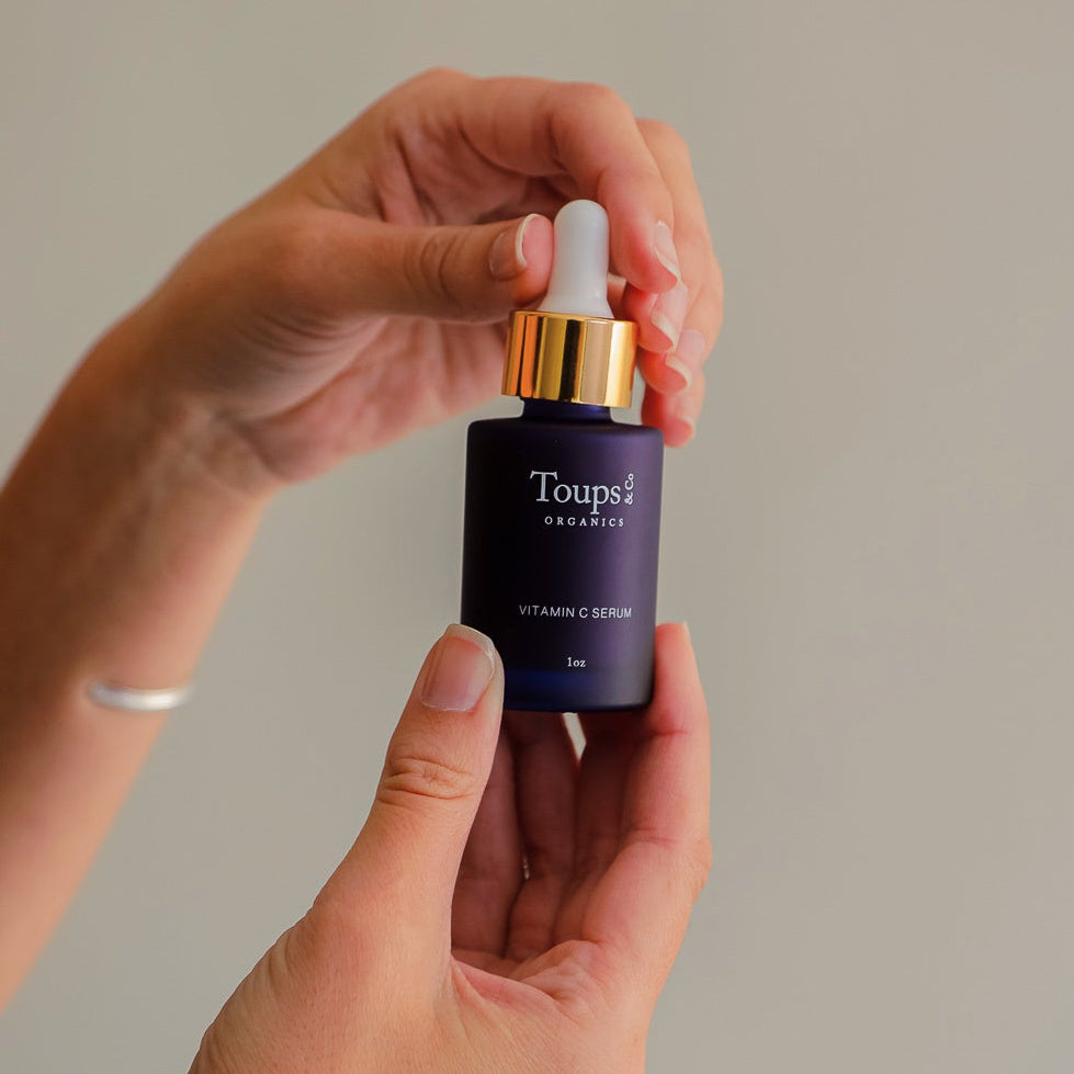 A person holds a dark glass dropper bottle labeled Toups and Co Organics Clean Serum Set against a plain light background.