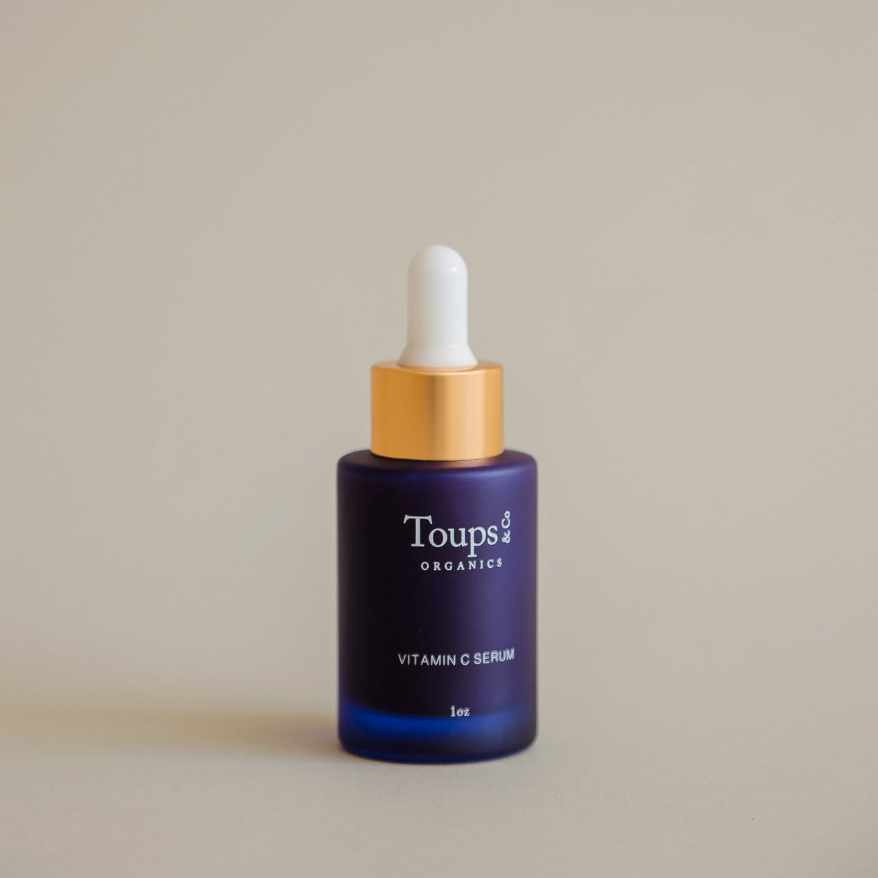 A blue glass dropper bottle labeled Toups and Co Organics Vitamin C Serum with a gold and white dropper cap, set against a plain beige background.