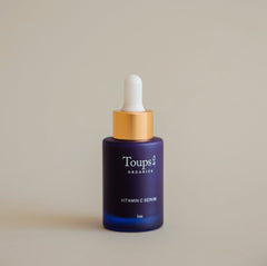 A blue glass dropper bottle labeled Toups and Co Organics Vitamin C Serum with a gold and white dropper cap, set against a plain beige background.