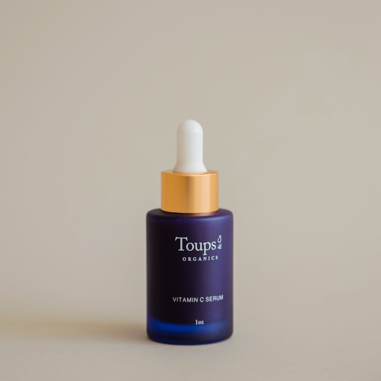 A blue glass bottle of Toups and Co Organics Vitamin C Serum with a gold and white dropper cap is shown against a plain beige background.