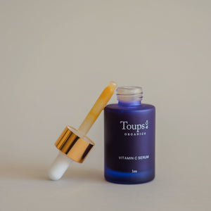 A blue glass bottle labeled Toups and Co Organics Vitamin C Serum is shown with a dropper filled with yellowish serum resting against it.