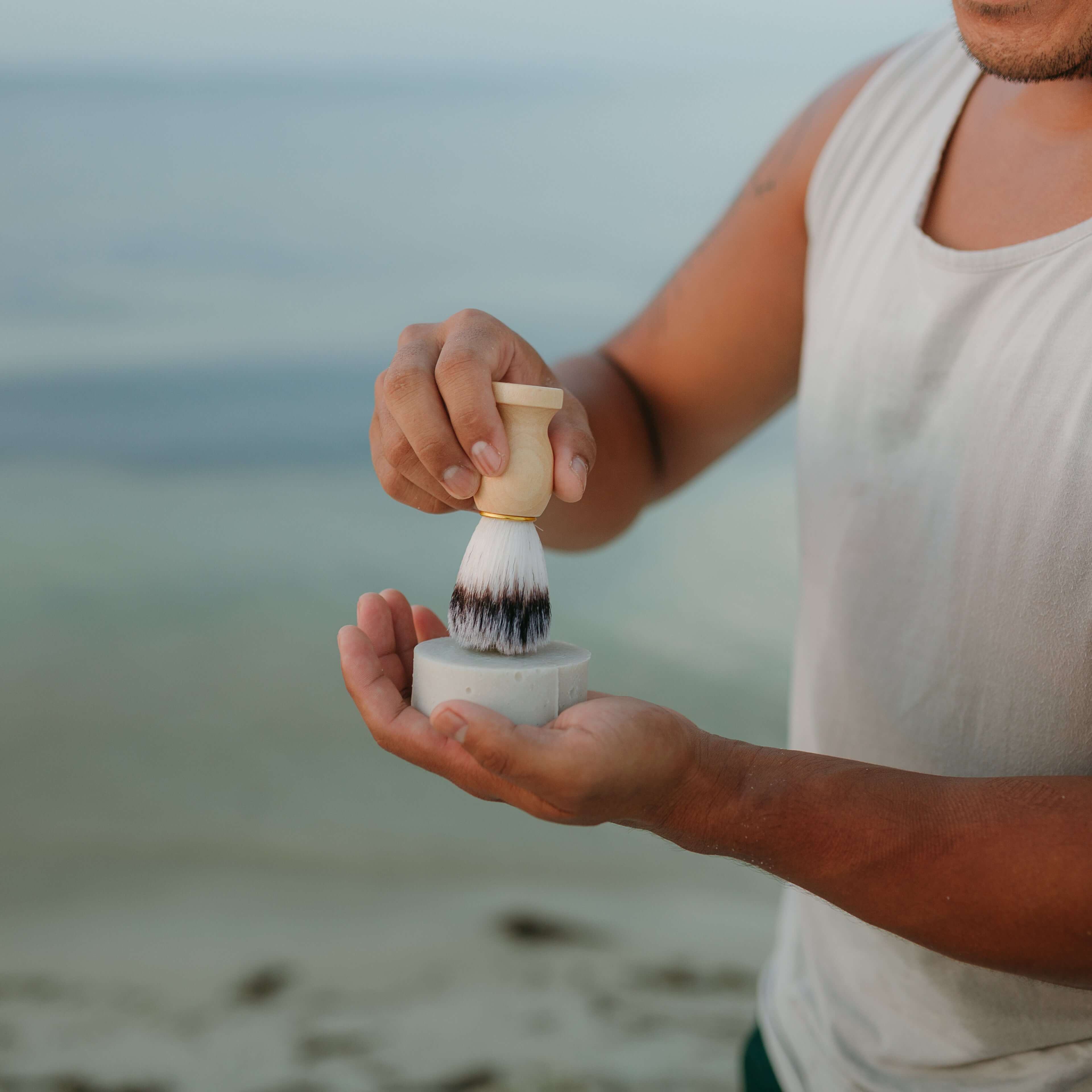 man-holds-up-tallow-shave-soap-and-shave-brush-against-beach-background